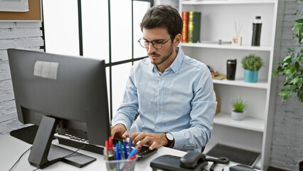 A focused hispanic man with a beard works at a computer in a modern office setting.