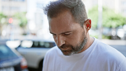 A mature hispanic man with grey hair looks down thoughtfully on a sunny urban street.