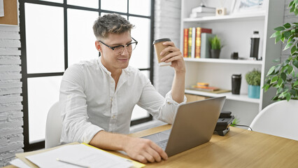 Fototapeta na wymiar Handsome young caucasian man, confident worker smiling at his desk in the office, enjoying morning espresso while working online with laptop
