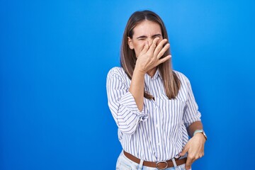 Hispanic young woman standing over blue background smelling something stinky and disgusting,...