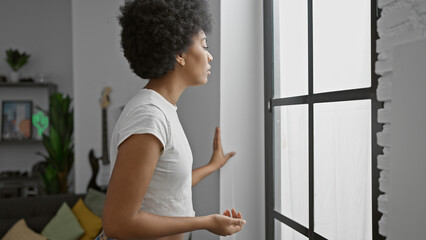 A contemplative african american woman with curly hair standing indoors near a window in a tidy...