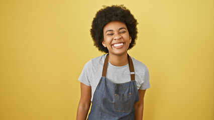A joyful african american woman with curly hair wearing a denim apron smiles against a yellow...