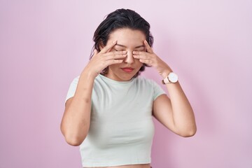 Hispanic young woman standing over pink background rubbing eyes for fatigue and headache, sleepy...