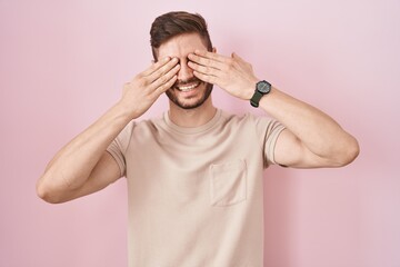 Hispanic man with beard standing over pink background covering eyes with hands smiling cheerful and...