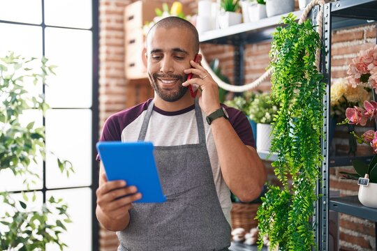 Young latin man florist talking on smartphone using touchpad at flower shop