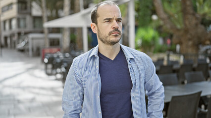 A pensive hispanic man with a beard and no hair stands on a city street with an outdoor cafÃ© background.