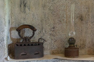 An old rusty charcoal iron and a kerosene lamp against the background of a cement wall.