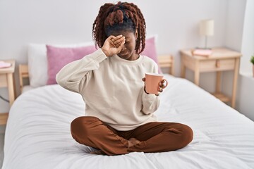 African american woman sitting on bed yawning and drinking coffee at bedroom