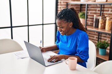 African american woman using laptop writing on document at home