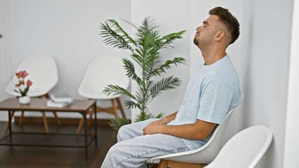 A young hispanic man relaxes in a modernly styled waiting room with white chairs and plants.