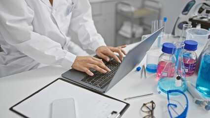 Hispanic woman scientist's hands deftly typing, immersed in groundbreaking medical research, working indoors at a bustling lab using laptop computer.