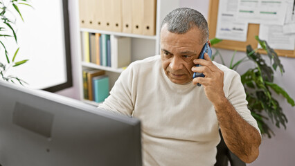 A mature hispanic man in a white sweater attentively talks on a smartphone in an office setting,...