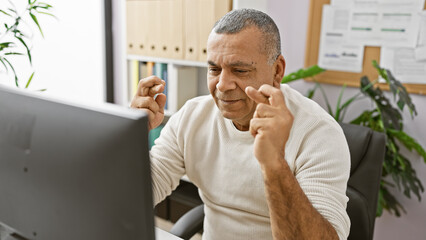 Middle-aged hispanic man crossing fingers for luck while working at his office computer indoors.