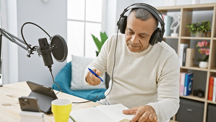 Mature hispanic man recording audio in a cozy, well-lit home studio, using a professional microphone and wearing headphones.