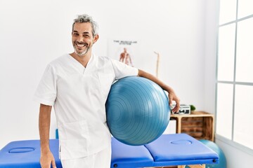 Middle age grey-haired man physiotherapist smiling confident holding yoga mat at rehab clinic