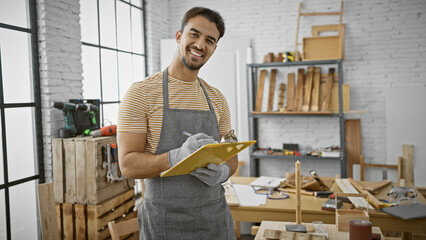 A handsome young hispanic man with a beard smiling in a carpentry workshop, wearing an apron and holding a clipboard.