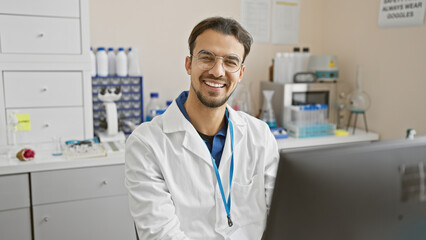 A smiling young hispanic man with a beard wearing glasses and a lab coat in a laboratory setting. - Powered by Adobe