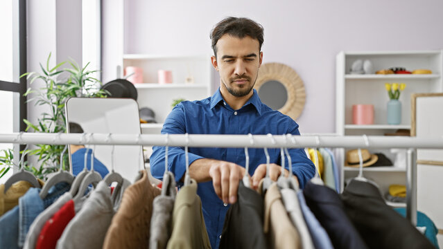 Handsome hispanic man with beard selecting clothes in a bright indoor room, reflecting fashion and wardrobe choices.