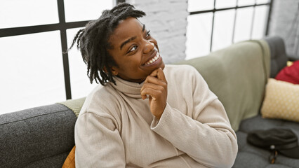 A smiling young black woman with dreadlocks wearing a turtleneck at home on a sofa.