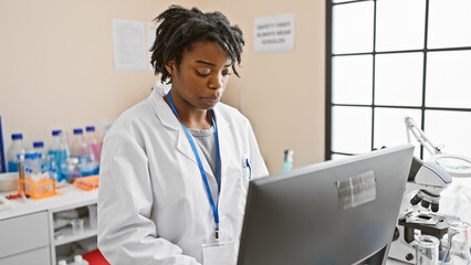 A focused african american woman in a lab coat analyzes data on a computer in a modern laboratory setting.