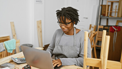 A young african american woman with dreadlocks working on a laptop in a carpentry workshop.