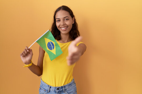 Young hispanic woman holding brazil flag smiling friendly offering handshake as greeting and welcoming. successful business.