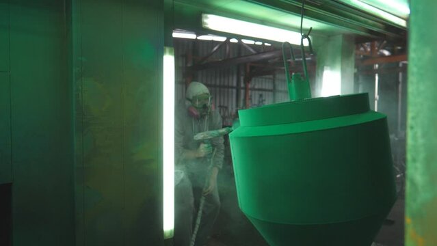Industrial painting process in chamber at factory. Painter spraying green paint on detail in special booth. Technician in safety wear working at production. Master paint metal in specialised workshop