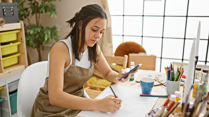 Enthralling glimpse of a beautiful young hispanic woman artist, earnestly drawing on her notebook, armed with a smartphone in hand at an art studio.