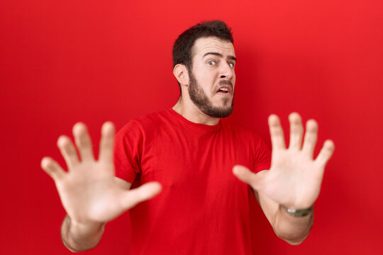 Young hispanic man wearing casual red t shirt afraid and terrified with fear expression stop gesture with hands, shouting in shock. panic concept.