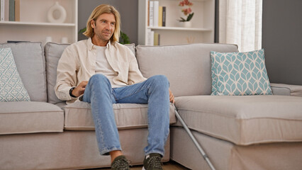 A relaxed man with long blond hair sitting on a modern sofa in a stylish living room, crutches nearby, implying injury.