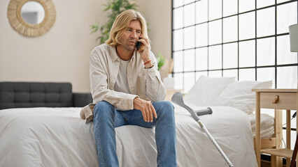 Blond man talks on phone sitting on bed with crutches in bright bedroom