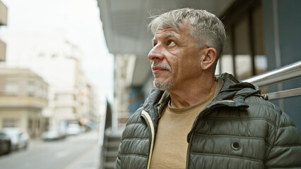 Mature grey-haired man in a jacket gazes thoughtfully outdoors in an urban street setting. - Powered by Adobe