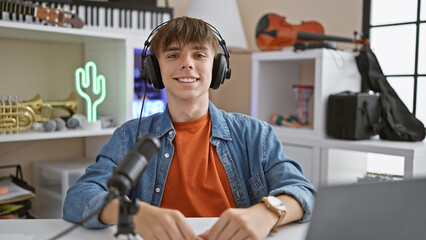 A cheerful young man in headphones sits in a modern room with musical instruments, engaging in a podcast or online streaming.