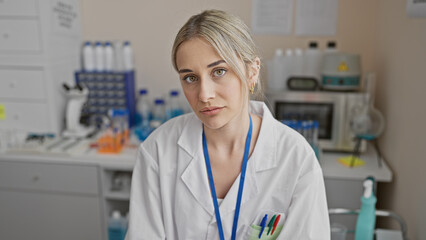 A young, blonde woman in a white lab coat poses thoughtfully in a laboratory, highlighting her...