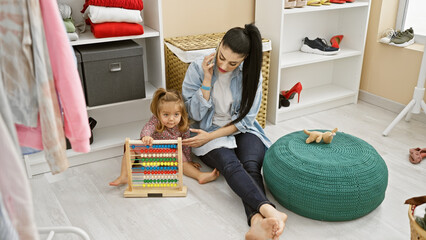 A woman talks on the phone while her daughter plays with an abacus in a cluttered dressing room.
