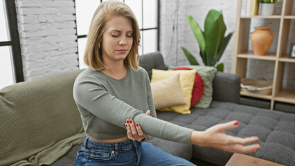 Young adult woman with short blonde hair touches her elbow in pain inside a cozy living room