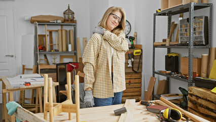 A beautiful young woman with short blonde hair wearing safety glasses experiences back pain in a woodworking workshop.