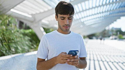 Cool looking young hispanic man, with serious expression, deeply engrossed in texting on smartphone...