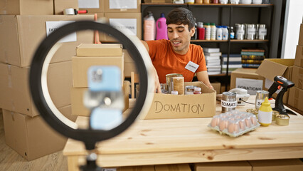 Radiant young hispanic man sitting at table, cheerfully volunteering at charity center, engaged in heartfelt video call via smartphone amidst stacked donation cardboard boxes