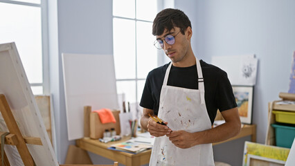 Attractive young hispanic man engrossed in his journey of creativity, holds paintbrushes in art...
