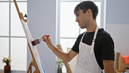 Young, handsome hispanic man engrossed in drawing, inside the vibrant art studio, the talented artist stands in apron, paintbrush in hand, studiously concentrating on his canvas