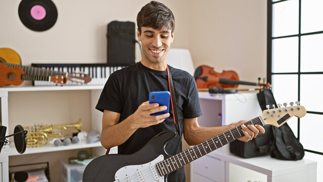 Attractive young hispanic man, an electric guitar musician, charmingly smiling while texting on his smartphone in a music studio, creating his own rock concert