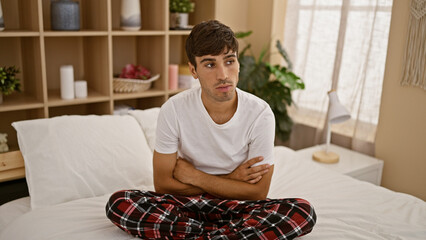 Upset young hispanic man, arms crossed, sitting on cozy bed looking serious in his bedroom...