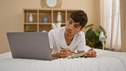 Handsome young hispanic man relaxing in bed, engrossed in writing into his notebook, laptop by his...