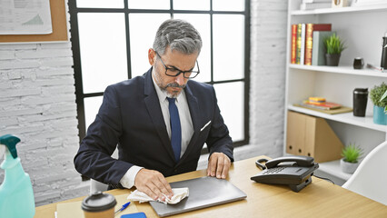 Handsome, grey-haired hispanic man cleaning his laptop at the office as part of his daily professional routine, exuding concentration and success at work