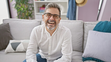 Cheerful young hispanic man with grey hair, sitting confidently on his living room sofa, letting...