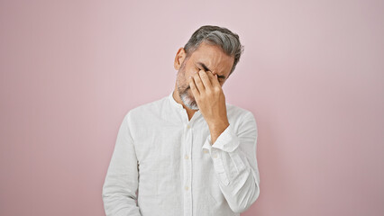 Young isolated hispanic man with grey-hair standing over pink background, visibly upset & frustrated expressing sadness & anxiety from unbearable headache ache, a symbol of mental stress and despair.