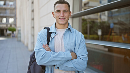Smiling young hispanic man with arms crossed wearing casual clothing on a sunny urban street.