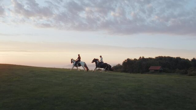 Two girls horseback riding in slowmotion at sunset. Horse galloping in the countryside on the top of the hill. Mountains and clouds in the background.