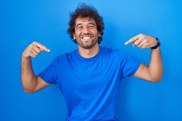Hispanic young man standing over blue background looking confident with smile on face, pointing...